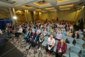 large number of people sitting in chairs in a big room, facing the camera