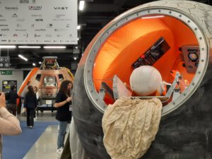 round orange opening of a space capsule and helmet of a dummy in foreground, model of an apollo capsule in the background